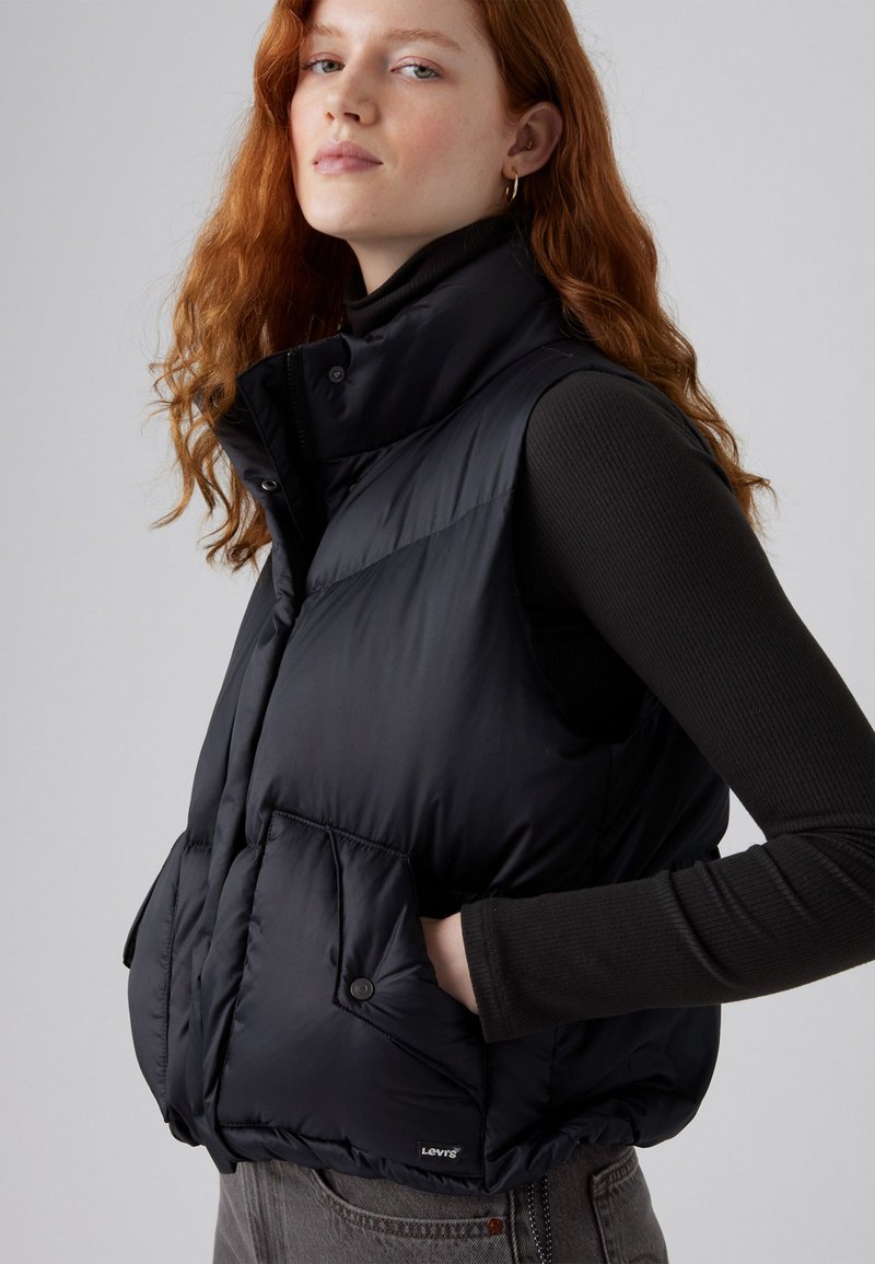 Gilet rembourré noir avec col haut, fermeture éclair et poches à bouton-pression. Présente des détails matelassés et un logo sur la poche gauche.