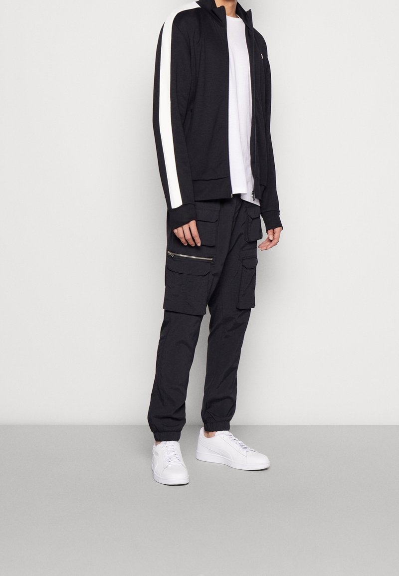 Homme portant une veste de survêtement noire avec des bandes blanches, un t-shirt blanc, un pantalon cargo noir avec des poches et des baskets blanches sur fond uni.