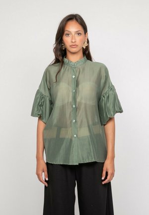Femme aux longs cheveux bruns portant une blouse boutonnée légère vert sauge avec des manches courtes froncées et un pantalon noir à jambes larges, debout devant un fond gris.