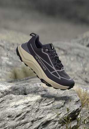 Čierno-béžová trailová bežecká topánka The North Face sa vznáša nad skalnatým terénom, ukazujúc robustnú podrážku a vonkajšie prostredie.