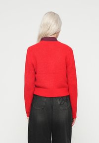 Punane kudumistega sviiter pikki varrukatega, ribatud servaga ja lõdva lõikega, kantud tumedate teksadega, mille tagakottidel on tikandid.