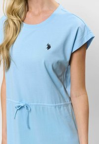 Šviesiai mėlynas medvilninis suknelė su apvaliu kaklu ir trumpomis rankovėmis. Yra nertinė liemuo ir mažas juodas logotipas ant krūtinės.