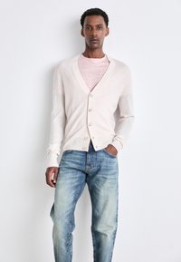 Gai beidzīgs kardigāns ar V-veida kakla izgriezumu, pogām priekšpusē un ribotām piedurknēm, uzvilkts virs rozā krekla, saskaņots ar izbalējušām zilu džinsiem.