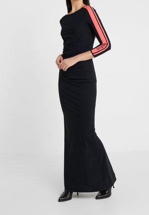 Femme portant une robe longue noire ajustée à manches longues avec une bande rouge et le texte "Patrizia" sur la manche gauche, associée à des chaussures noires pointues.