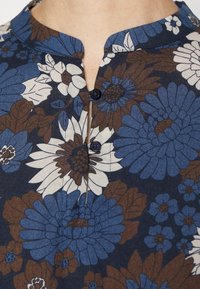 Top con motivo floreale nei toni di blu e marrone, con colletto rotondo e tre bottoni scuri all'altezza dello scollo su uno sfondo scuro.