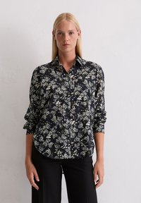 Femme aux cheveux blonds raides portant une chemise à boutons à manches longues florale noire et un pantalon noir, se tenant contre un mur blanc.