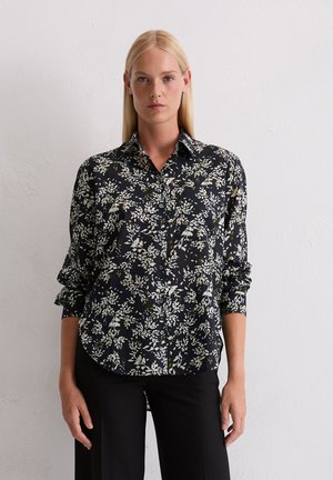 Femme aux cheveux blonds raides portant une chemise à boutons à manches longues florale noire et un pantalon noir, se tenant contre un mur blanc.