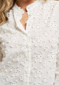 Blouse blanche brodée avec des motifs floraux, des coutures texturées, des boutons sur le devant et un col mandarin.
