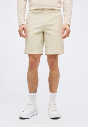 Mees, kes kannab beeže lühikesi pükse, valget pika varrukaga pluusi, valgeid sokke ja valgeid tossusid, seisab käed vabalt külgedel.