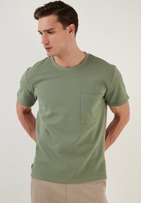 T-shirt verde oliva a maniche corte con collo rotondo, tessuto testurizzato e una tasca sul petto sinistro. Taglio dritto sull'orlo.