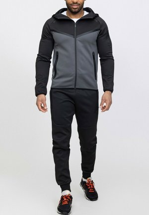 Ensemble de vêtements de sport comprenant une veste zippée à capuche grise et noire avec deux poches avant, accompagnée de pantalons noirs fuselés et de baskets noires.