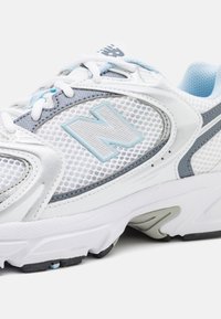 Weißer Sportschuh mit Mesh-Obermaterial, hellblauen Akzenten und grauen Details. Verfügt über eine gepolsterte Sohle und ein strukturiertes Profil für besseren Grip.