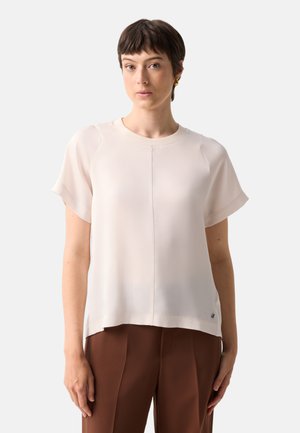 Kvinde med kort brunt hår iført en ensfarvet, lys beige kortærmet top og brune bukser mod en hvid baggrund.