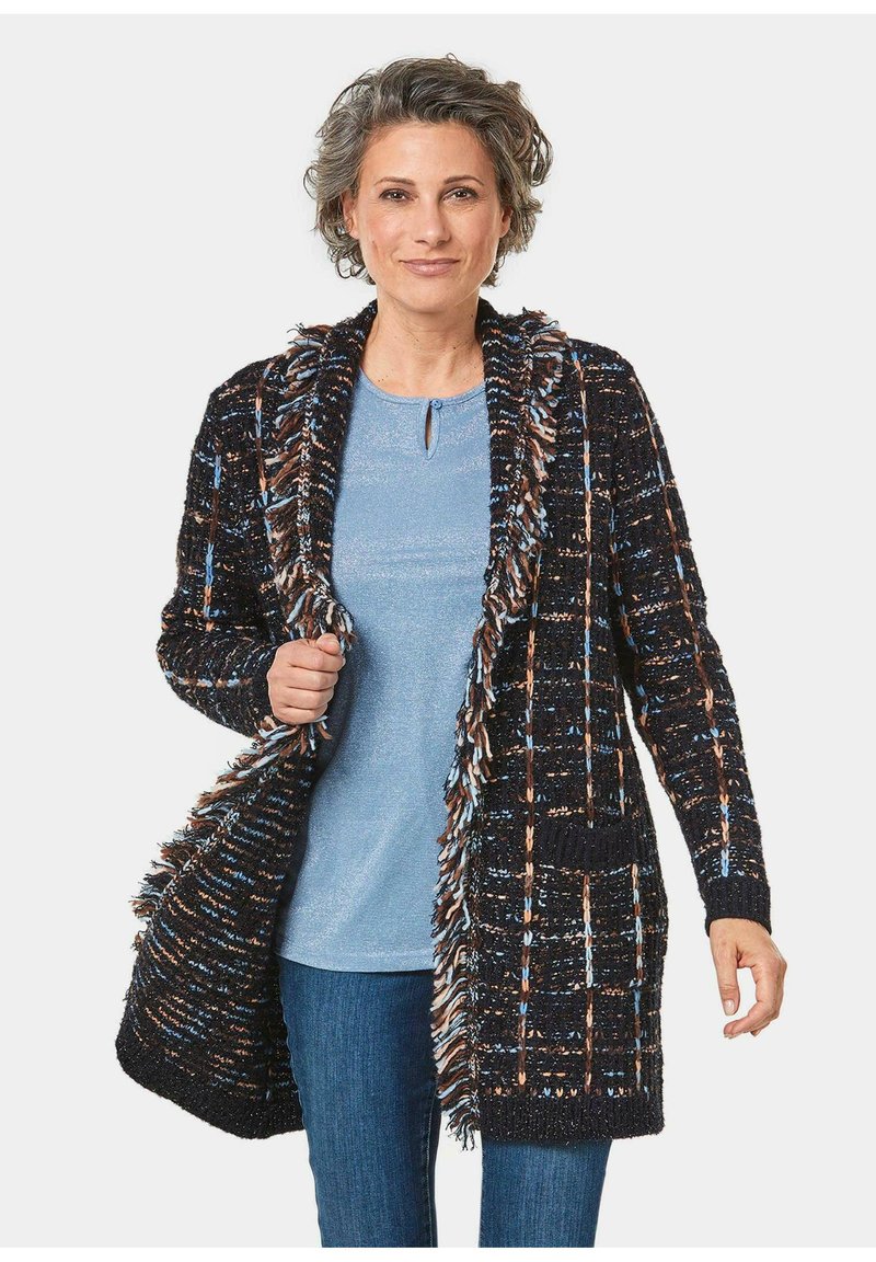 Schwarze und mehrfarbige Strickjacke mit einer Fransenkragen, offener Front und zwei Taschen. Getragen über einem blauen Oberteil, kombiniert mit blauen Jeans.