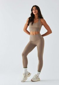 Beżowy zestaw sportowy składający się z krótko skrojonego topu na szerokich ramiączkach oraz legginsów z wysokim stanem. Modelka ma na sobie białe sneakersy z zielonymi akcentami.