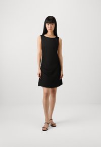 Vestido preto sem mangas com gola redonda, corte solto e detalhe de drapeado lateral. Modelo usando sapatos de salto pretos com tiras.