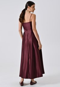 Robe maxi en satin bourgogne avec de fines bretelles, un corsage ajusté et une jupe fluide. Dotée d'une fermeture à glissière au dos et d'une texture lisse.