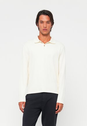 Camisa polo de manga larga, color crema, de tejido suave al tacto, con cuello abotonado, dobladillo recto, combinada con pantalones oscuros.
