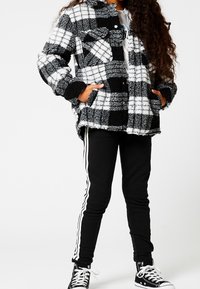 Chaqueta de forro polar a cuadros en blanco y negro con dos bolsillos, cierre de botón y una textura suave, acompañada de pantalones negros con rayas blancas.