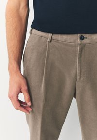 Pantalon beige en mélange de coton avec une texture lisse, doté d'une fermeture à bouton unique et de plis subtils. Porté avec un haut bleu foncé.