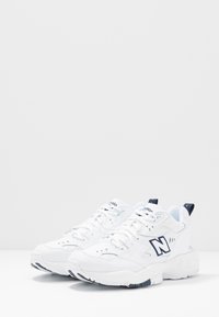 Bílé sportovní boty s vršky z síťoviny a kůže, polstrovaným límcem, šněrováním a tmavě modrými akcenty. Silná texturovaná bílá podrážka.