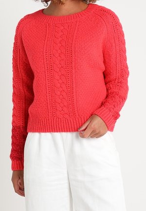Femme portant un pull en torsades rouge vif à manches longues et un pantalon blanc, debout devant un fond uni.