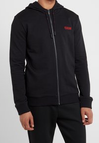 Felpa nera con zip realizzata in tessuto morbido, dotata di cappuccio, logo rosso sul petto e polsini ed orlo elasticizzati per una vestibilità aderente.