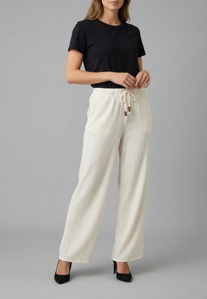 Donna con top nero a maniche corte, pantaloni bianchi a gamba larga con coulisse e perline di legno, e scarpe nere con tacco alto su sfondo grigio.