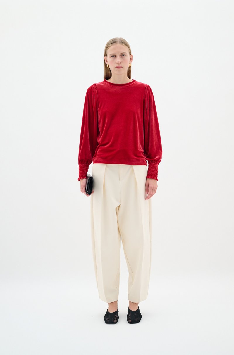 Rotes Samt-Oberteil mit langen Ärmeln und gerafften Bündchen, kombiniert mit cremefarbenen, übergroßen Hosen und schwarzen Netzschuhen. Das Model hält eine kleine schwarze Clutch.