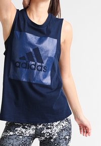 Sininen ilmanhihoinen t-paita, jossa on kiiltävä musta Adidas-logo edessä. Yhdistetty kuosisin legginsien kanssa, joissa on pikselimäinen kuvio harmaassa ja valkoisessa.