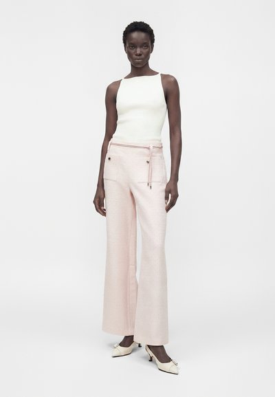 Γυναίκα που στέκεται, φοράει λευκό αμάνικο τοπ, ανοιχτό ροζ φαρδιά παντελόνια με τσέπες μπροστά και κρεμ παπούτσια με χαμηλό τακούνι και φιόγκους.