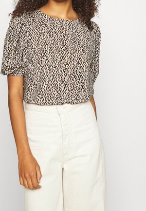 Kobieta w krótkim rękawie, nosząca bluzkę w cętki lamparta, włożoną do wysokich, kremowych spodni, na jednolitym tle.