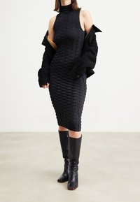 Černé bezramínkové šaty s texturovaným, prošitým vzorem a vysokým výstřihem, doplněné černou oversize bundičkou a černými botami po kolena.