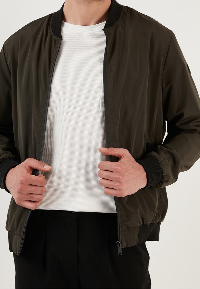 Oliivivihreä pommitakkia, jossa on sileä pinta, röyhelöhaalarit ja kaulus, etuvetoketju ja sivutaskut, joka on puettu valkoisen röyhelöpaidanne päälle.