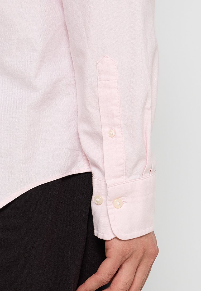 Eine Nahaufnahme einer Hand, die ein hellrosa Hemd trägt und das Kissen mit Knöpfen zeigt, vor einem schwarzen Hintergrund.