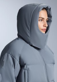 Sivi kaput od pufanja s velikim kapuljačom, ima glatku teksturu i vidljive šavove. Uključuje RECCO oznaku na rukavu.