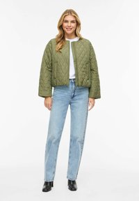 Moteris vilki alyvuogių žalios spalvos siuvinėtą striukę, baltą marškinius, šviesiai mėlynas džinsus ir juodus aukštakulnius batai iki čiurnos, stovinti prieš baltą foną.