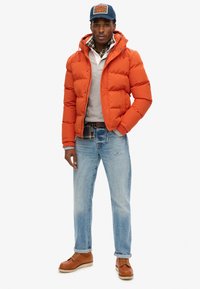 Orange pufferjakke, letvægts med lynlås og snoretræk i hætten. Grå sweater, lyseblå jeans, brune snørebåndsstøvler, marineblå kasket.