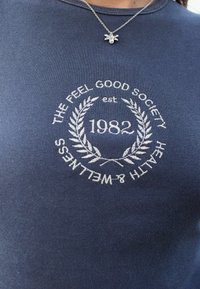 Tumesinine T-särk ribikangasest, millel on valgest tikandist tekst ja lauri disain. Tekst sisaldab "THE FEEL GOOD SOCIETY" ja "1982."