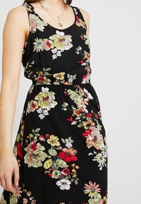 Czarna sukienka bez rękawów z elastycznym pasem, ozdobiona dużymi kwiatowymi wzorami w odcieniach czerwieni, żółci, bieli i zieleni, noszona z warstwami złotych naszyjników.