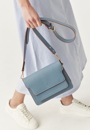 Geantă crossbody din piele albastră cu clapetă pliabilă, cusături accentuate maro și o curea de umăr detașabilă, ținută de o mână.