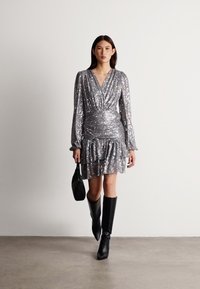 Hõbedane sädelev kleit, millel on tõmblukuga disain, pikad varrukad ja voltidega äär, koos põlvikute pikkuste mustade saapade ja väikese musta käekotiga.