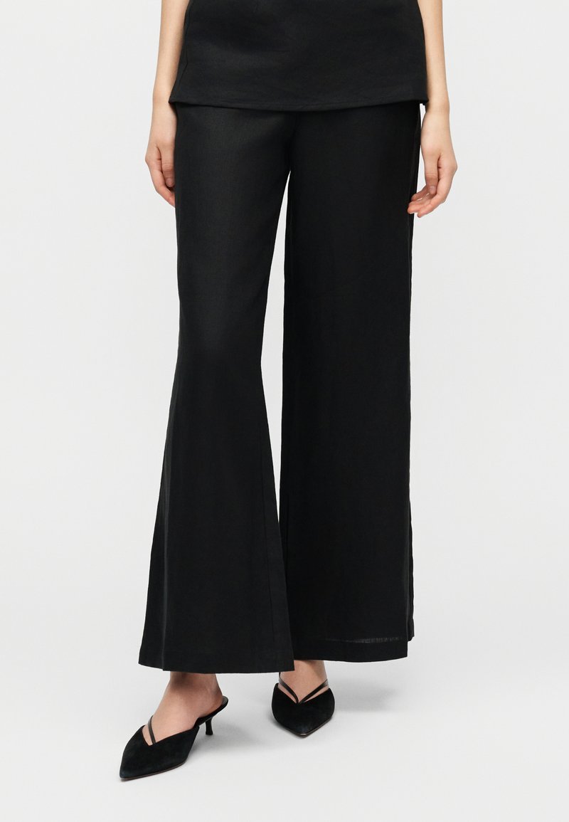 Μαύρο παντελόνι με καμπάνα από λεί ο ύφασμα, με ψηλή μέση και φαρδιές ποδιές, συνδυασμένο με μυτερά μαύρα παπούτσια χωρίς κορδόνια.