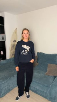 Camisola azul-marinho com um gráfico de peixe, calças escuras e ténis pretos. No fundo, um sofá azul-petróleo e uma prancha de surf.