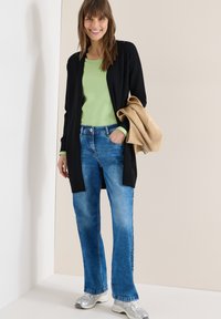 Femme souriante en jean bleu, haut vert, cardigan noir, tenant une veste beige, debout contre des murs beige et blanc, portant des baskets argentées.