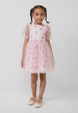 Jong meisje met gevlochten haar, gekleed in een roze jurk met aardbeienpatronen en pofmouwen, staande tegen een effen achtergrond.