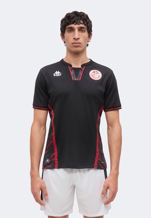 Jeune homme debout face à l’objectif portant un maillot de football noir à accents rouges et un short blanc, avec les logos Kappa et de la fédération de football tunisienne.