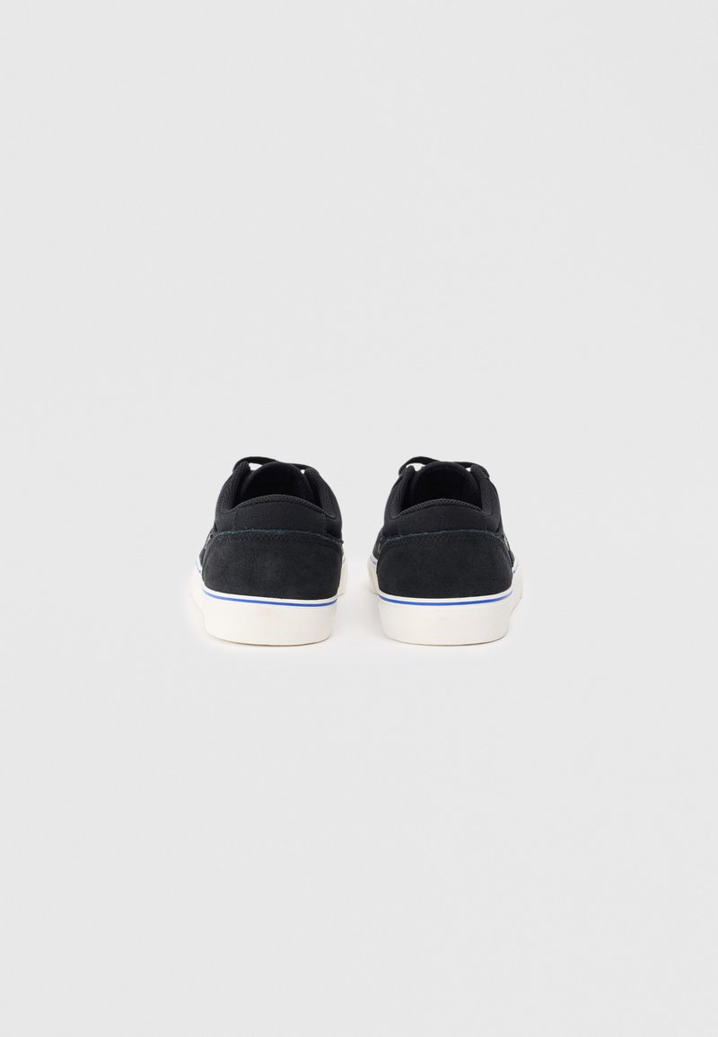Czarne zamszowe trampki z białymi gumowymi podeszwami, z przeszywanym wzorem i niebieskimi akcentami wzdłuż krawędzi. Widok z tyłu pokazujący podwójne uchwyty.