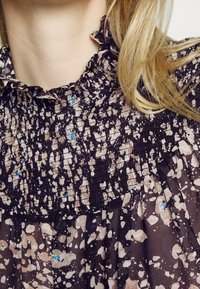 Blouse à motifs floraux sombres, avec un col volanté, un corsage froncé, et un motif de taches claires et foncées avec des touches de bleu.