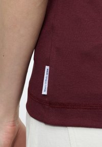 Κοντινό πλάνο στο χέρι ενός ατόμου δίπλα στο τελείωμα μιας μπλούζας μπορντό πλεκτής με ετικέτα υφάσματος «Marc O'Polo DENIM» και στη ζώνη μέσης λευκού παντελονιού.